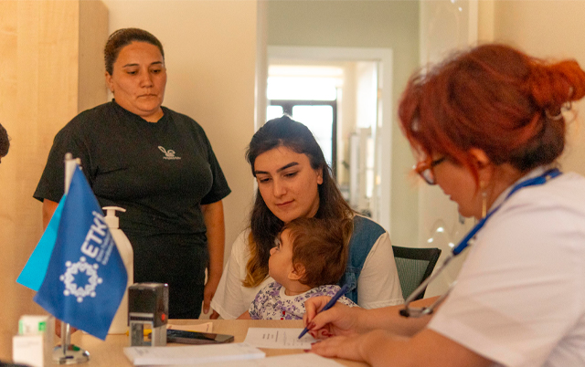 A healthcare setting with a nurse attending to a mother and child, while another woman observes. Medical paperwork is on the table.