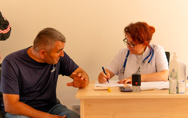 A doctor in a white coat takes notes while talking to a patient in a casual setting.