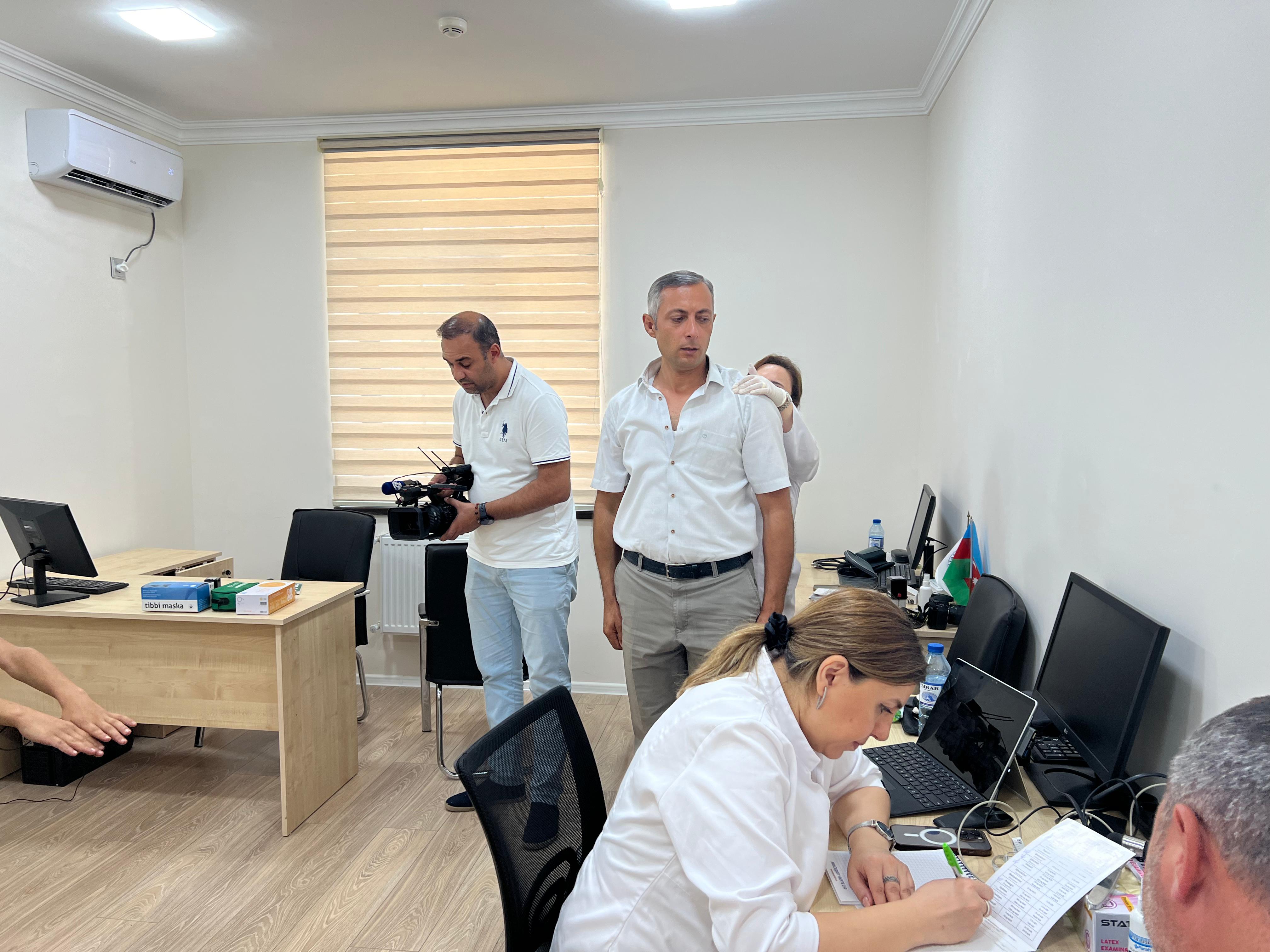 A group of people in an office setting, with one person filming, another writing notes, and two others standing nearby.