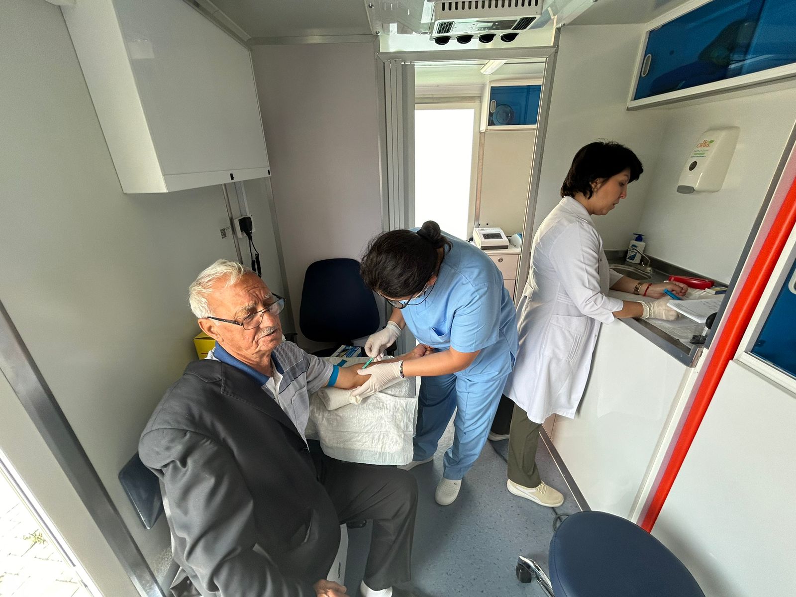 An elderly man sits in a medical setting while a nurse prepares to take his blood. Another staff member is at a counter.