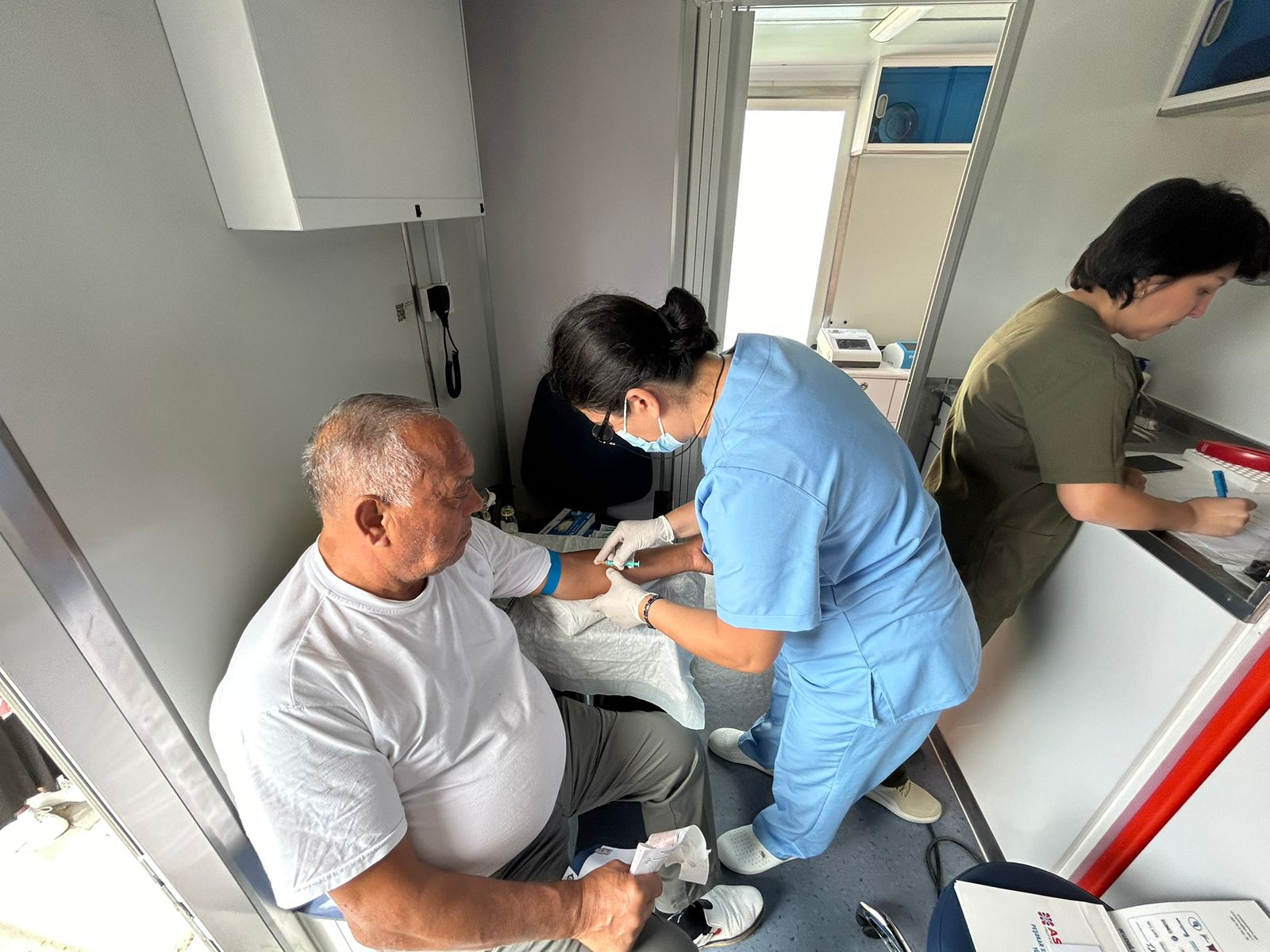 A healthcare worker is drawing blood from an elderly man in a medical setting, while another staff member is writing notes.