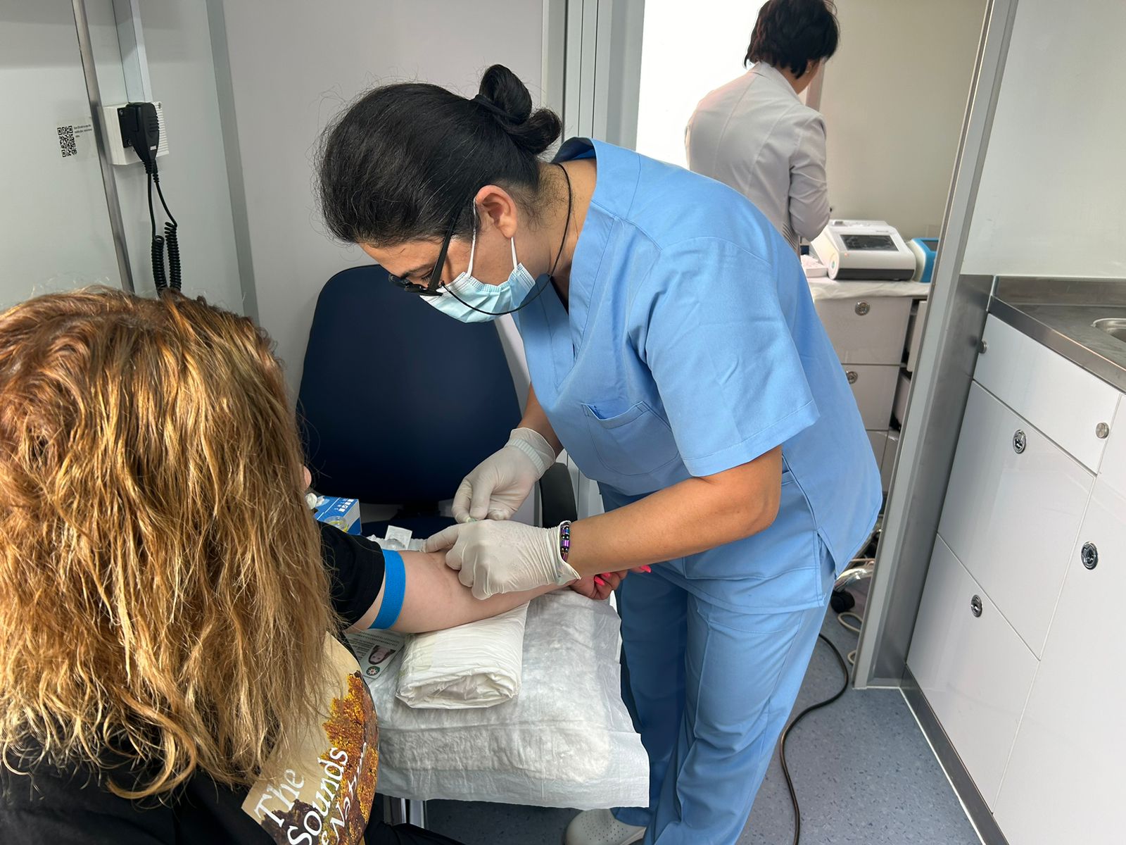 A healthcare professional in blue scrubs is drawing blood from a patient with curly hair in a medical setting.