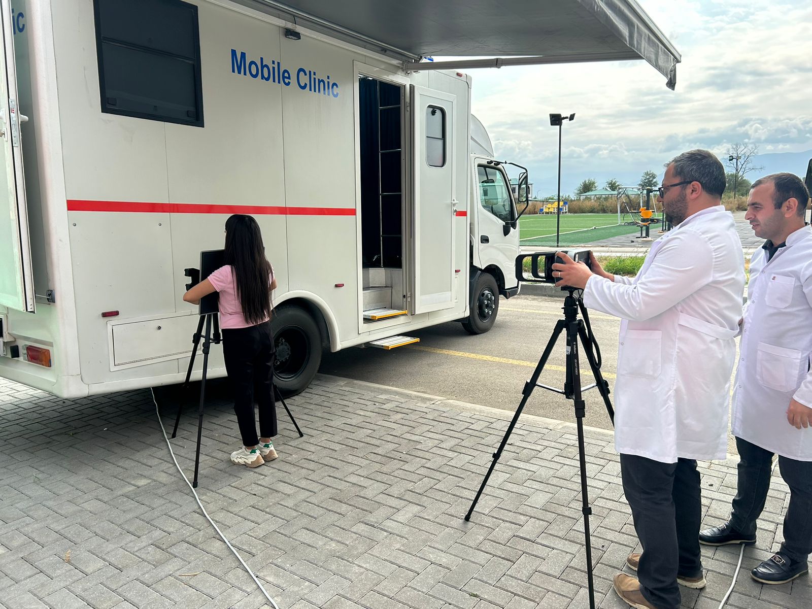 A mobile clinic is parked, with a girl using a camera on a tripod and two men in lab coats observing.