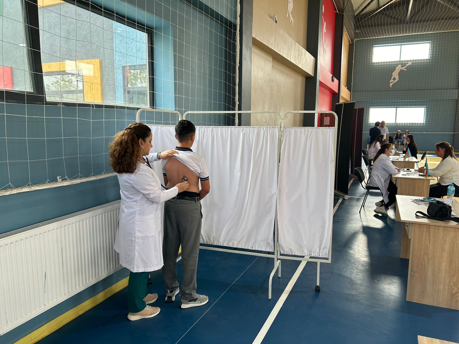 A healthcare professional examines a child's back with a stethoscope in a gymnasium setting, with other medical staff in the background.