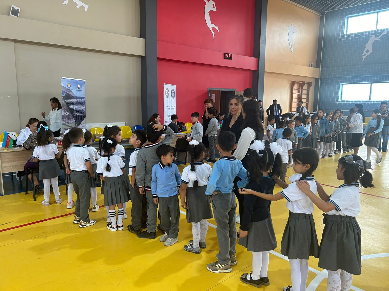 A group of children in school uniforms is lined up in a gymnasium, with adults supervising and a registration table in the background.