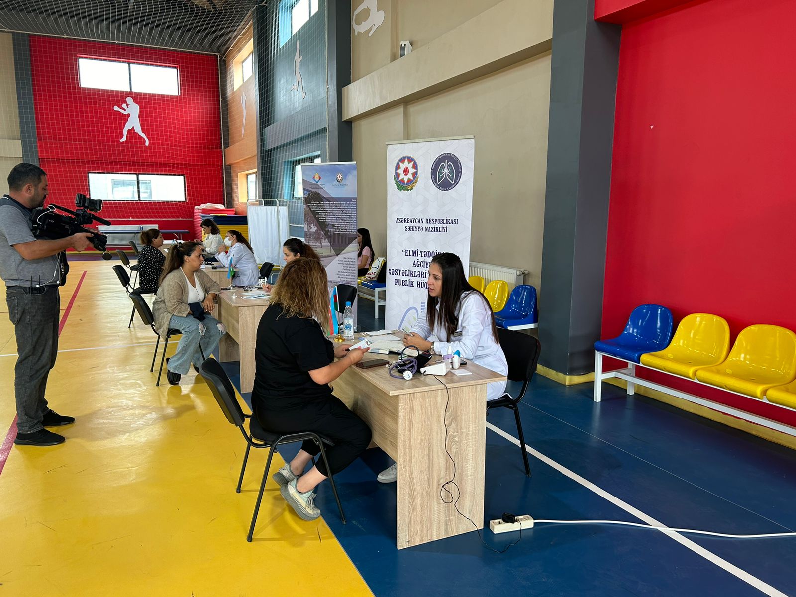 A health event in a gymnasium with medical staff assisting individuals at tables, while a cameraman records the activities.