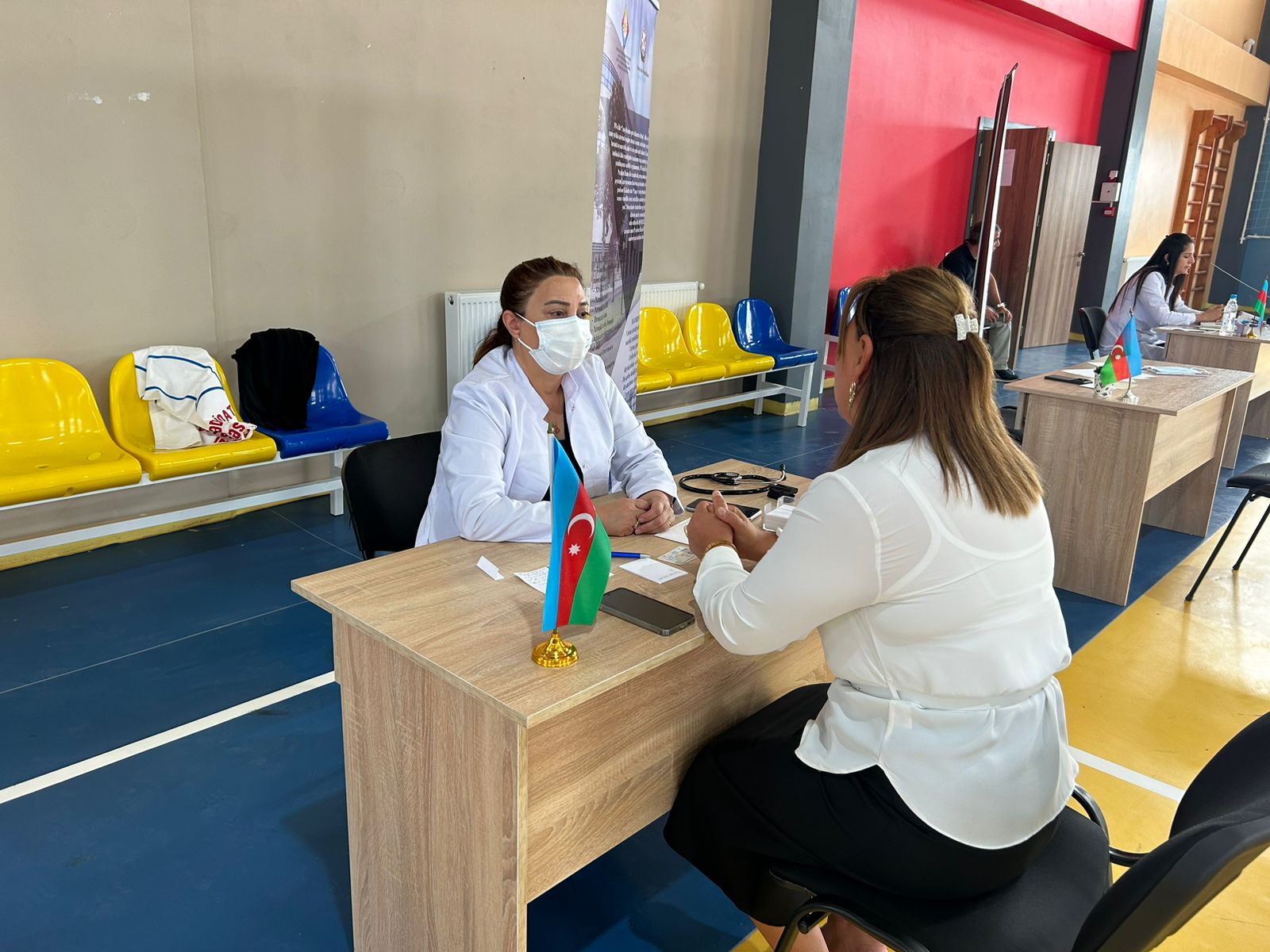 A woman in a white coat and mask sits at a table with an Azerbaijani flag, talking to another woman in a white blouse.