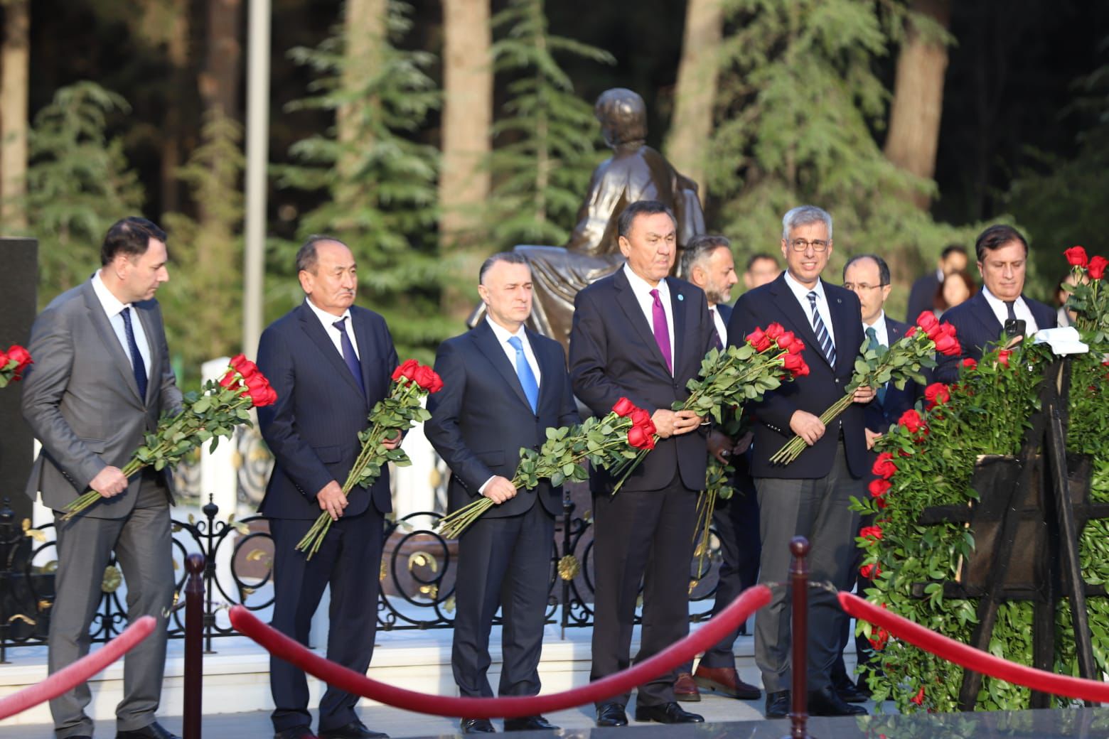 A group of men in suits holding red roses stands solemnly at a memorial site, surrounded by greenery.