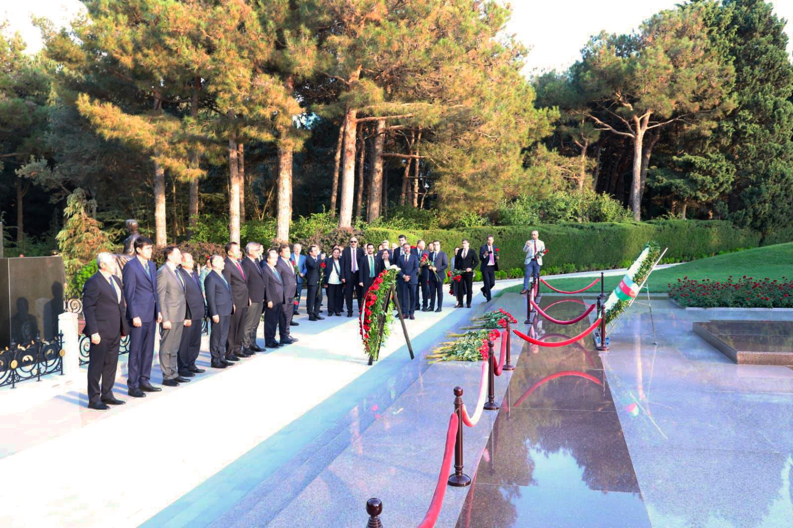 A group of officials stands in a memorial park, paying respects with floral wreaths, surrounded by trees and greenery.