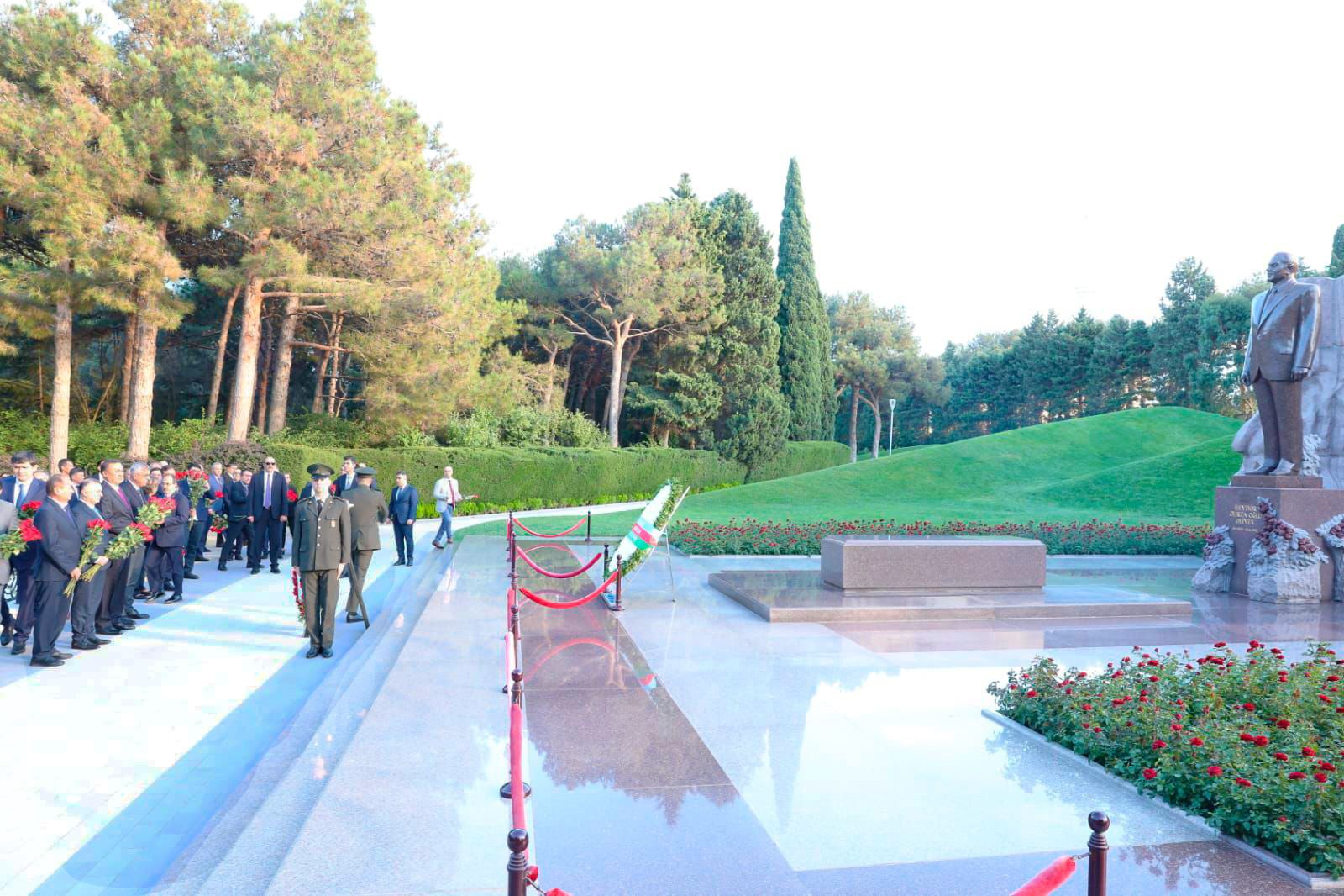 A group of people in formal attire stands near a monument, holding red flowers, in a landscaped area with trees and flowers.