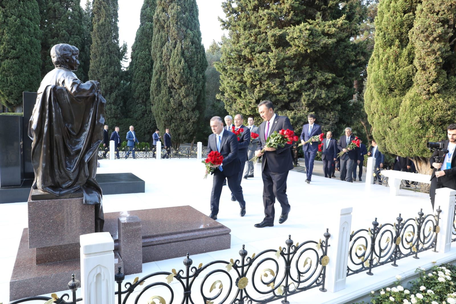 A group of officials in suits carries red roses while walking towards a statue in a memorial park surrounded by trees.