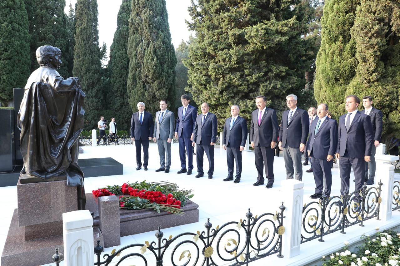 A group of officials stands in front of a statue, with a floral tribute of red roses nearby, surrounded by trees.