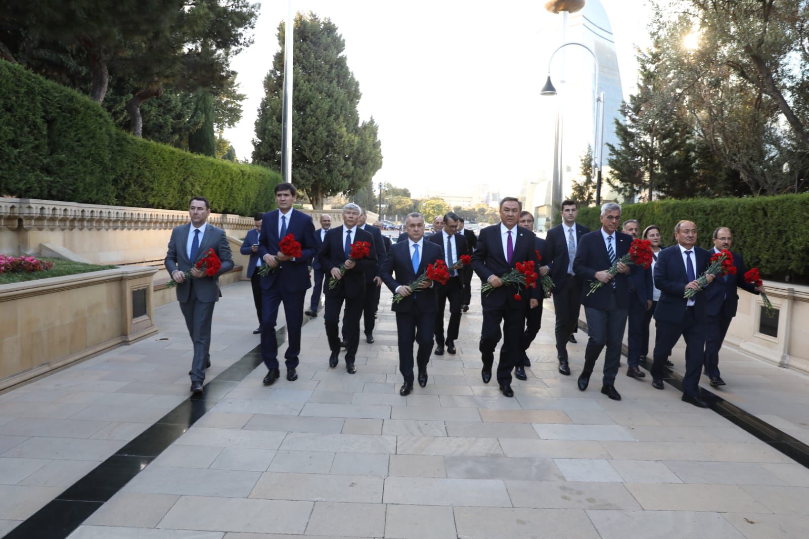 A group of men in suits walking together, holding red flowers, in a landscaped outdoor setting.