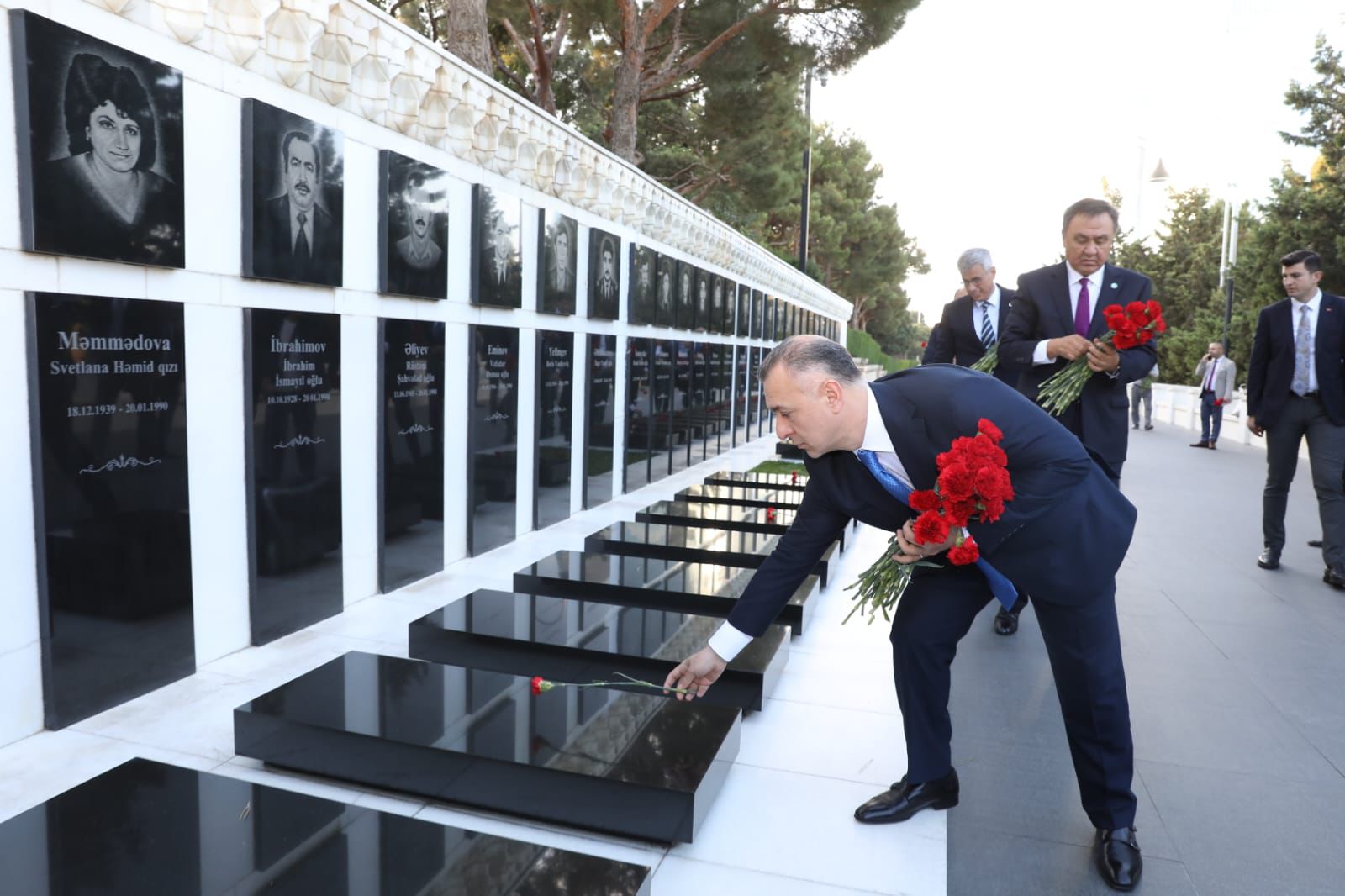 A group of men in suits lay red flowers at a memorial with black plaques, honoring individuals. Trees line the background.