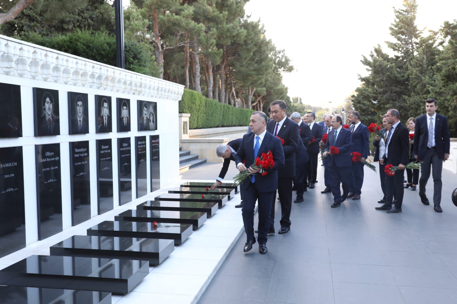 A group of people in formal attire are laying red flowers at a memorial wall with portraits, surrounded by greenery.