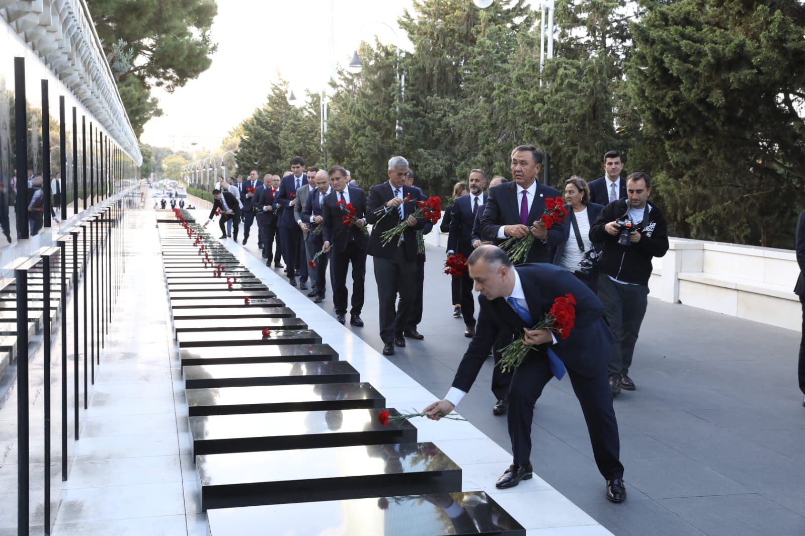 A group of people in formal attire are laying red flowers at a memorial site, surrounded by trees and reflective panels.