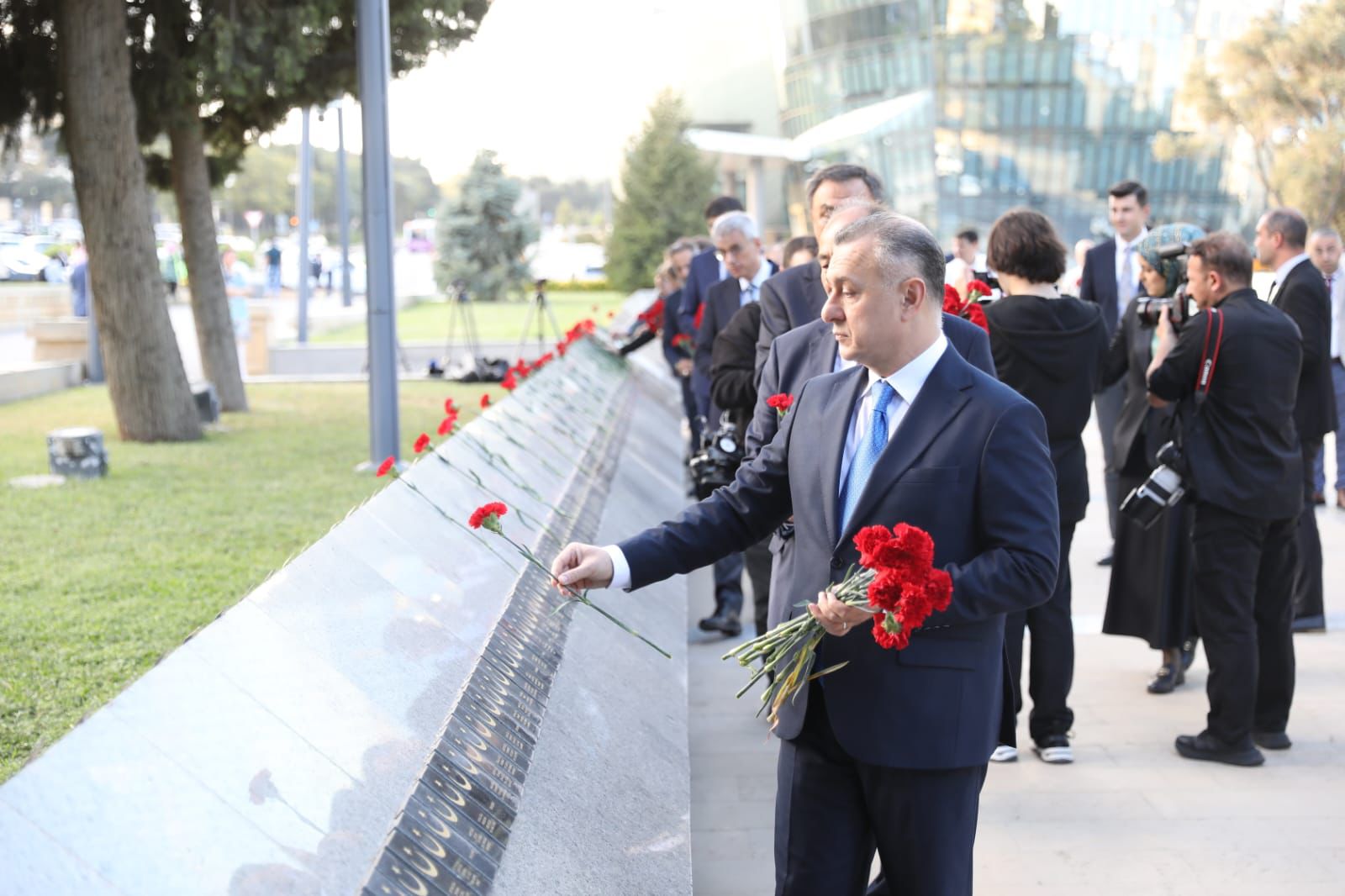 A group of people, including a man in a suit, are placing red carnations at a memorial while others observe and take photos.