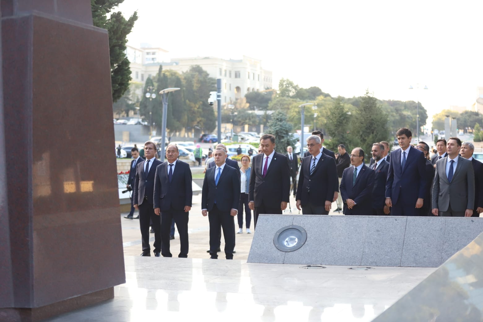 A group of men in suits stands solemnly in front of a monument, with trees and buildings in the background.