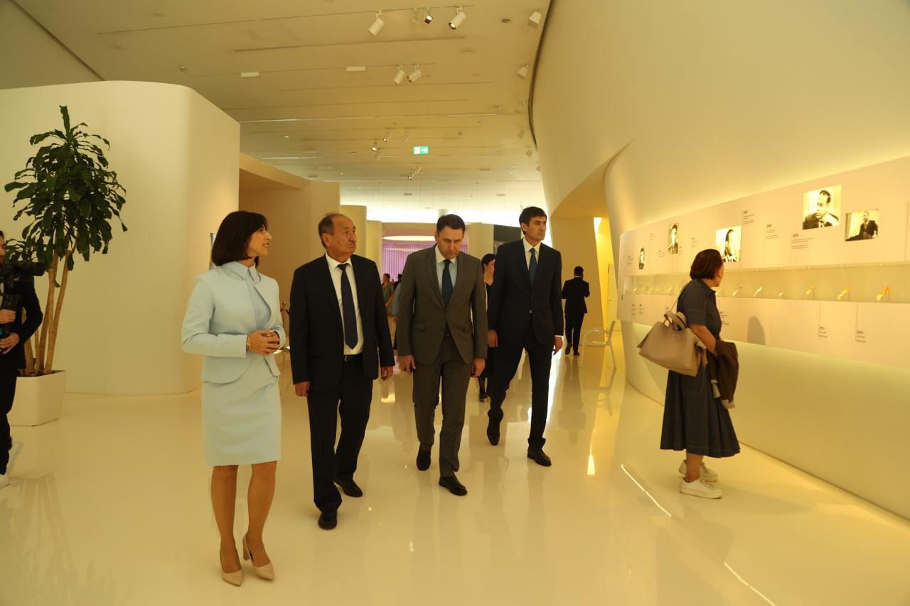 A group of people walks through a modern exhibition space, observing displays on the walls. A plant is visible in the background.