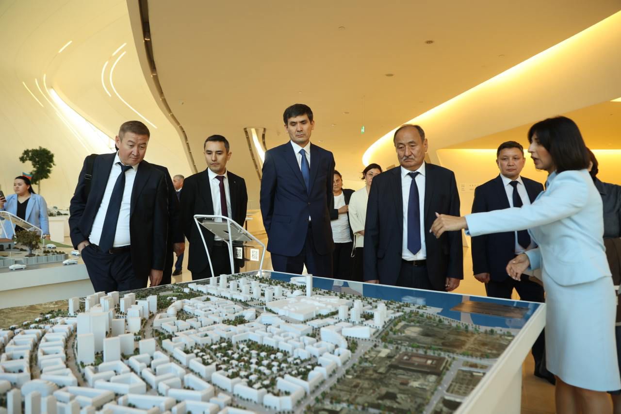 A group of professionals observes a city model while a woman explains details, set in a modern interior space.