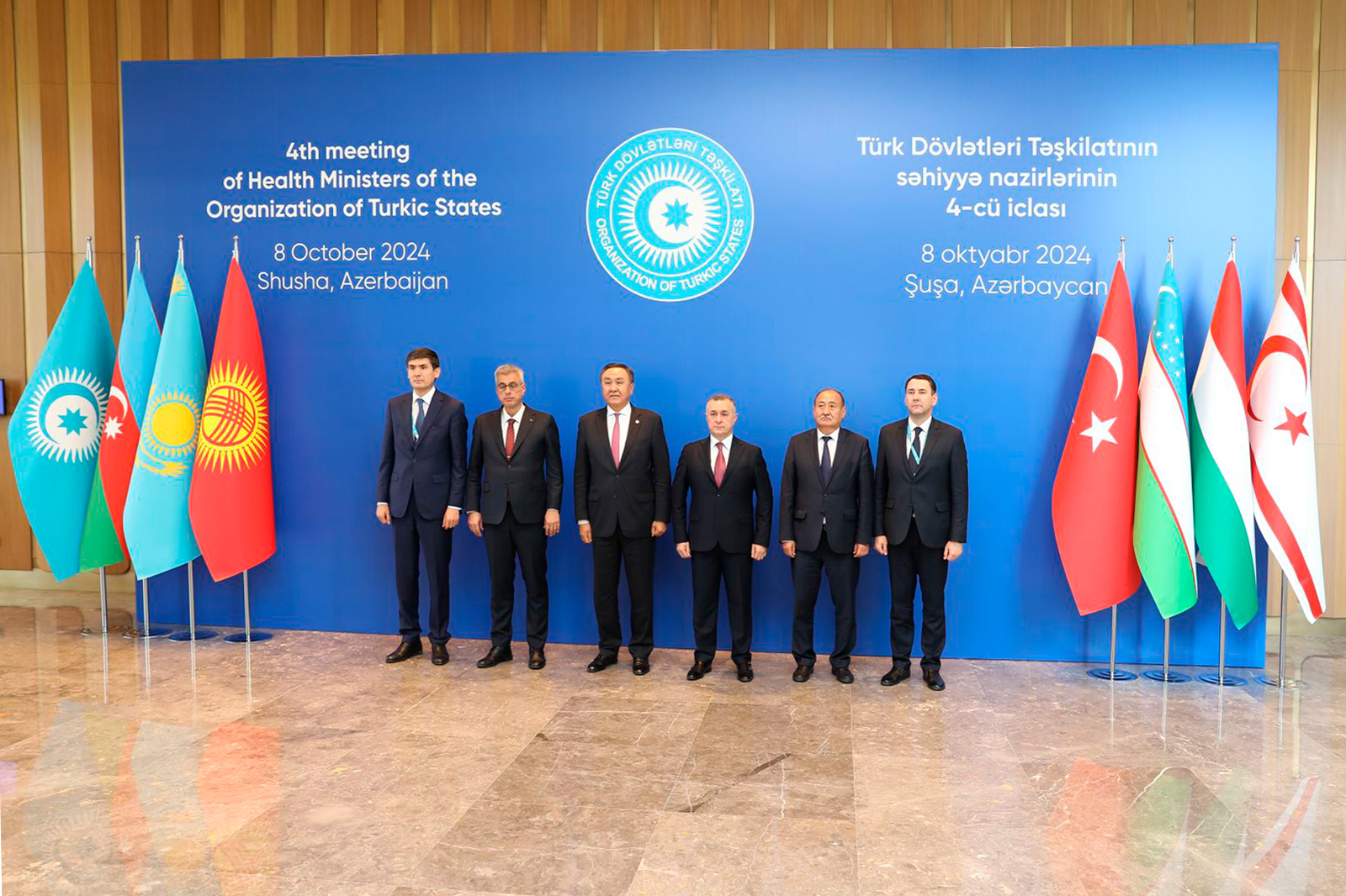 A group of officials stands in front of a blue backdrop with flags, marking the 4th meeting of Health Ministers of the Organization of Turkic States.