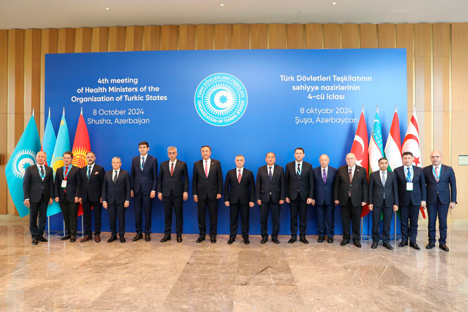 A group of officials stands in front of a blue backdrop, featuring flags of Turkic states, at a health ministers' meeting in Shusha, Azerbaijan.