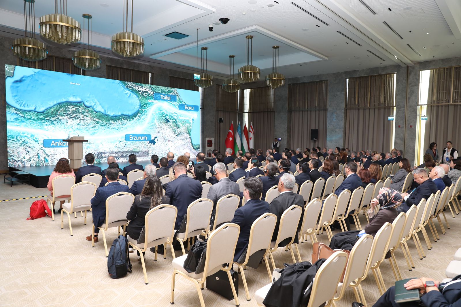 A conference room filled with attendees facing a large screen displaying a map of the region, with flags in the background.