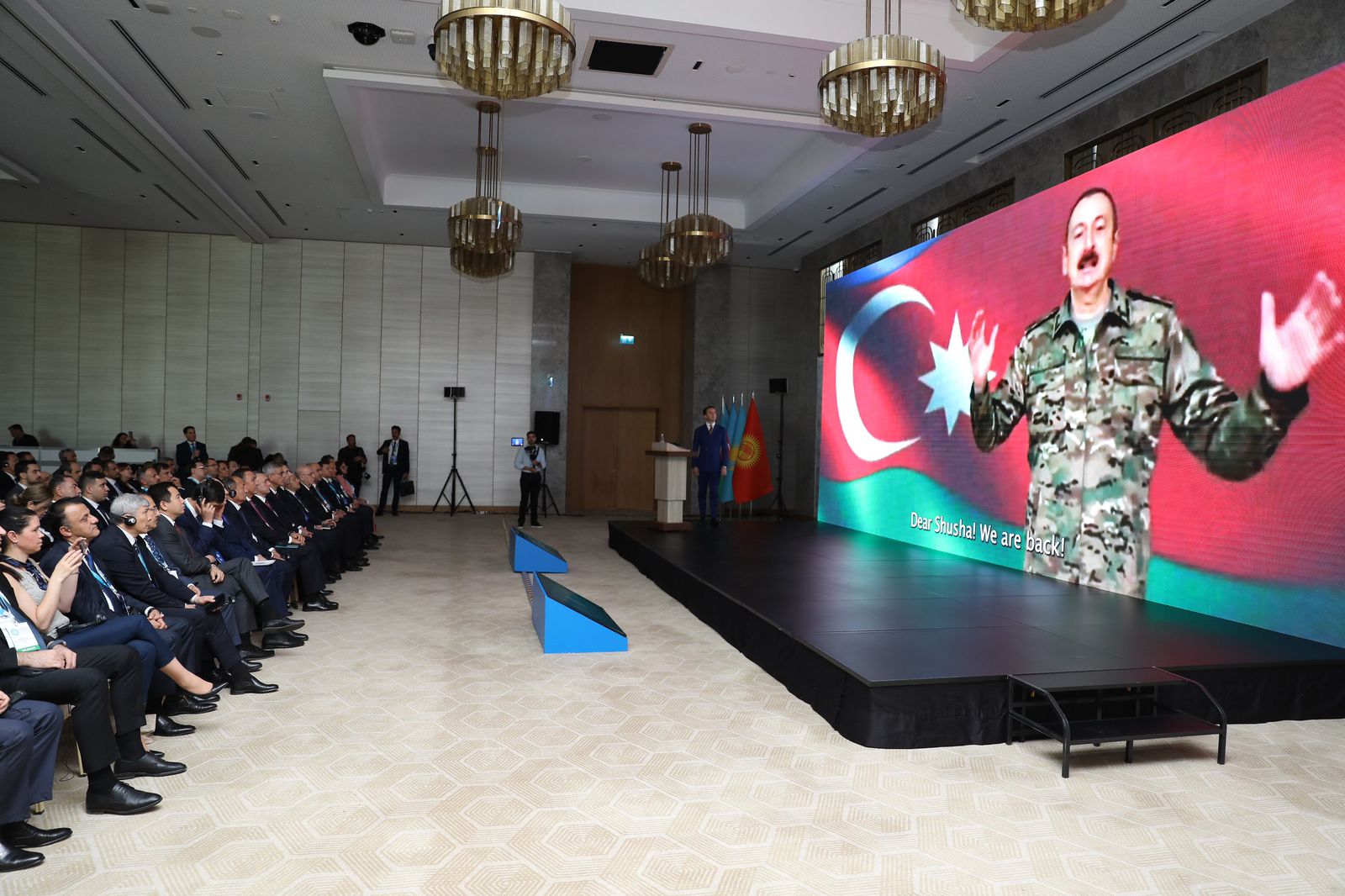 A large audience watches a presentation featuring a speaker in military attire against a backdrop of flags and a digital screen.