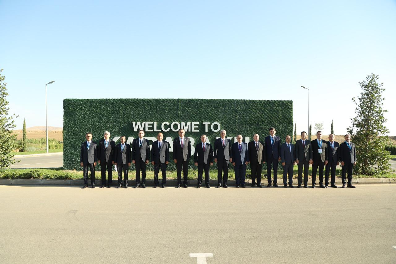 A group of men in suits stands in front of a large "WELCOME TO" sign, with greenery and a clear blue sky in the background.