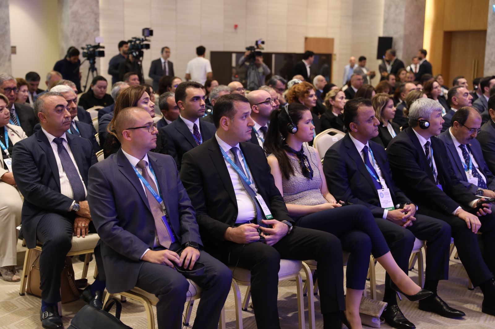 A diverse audience seated in a conference hall, attentively listening, with some wearing headphones.