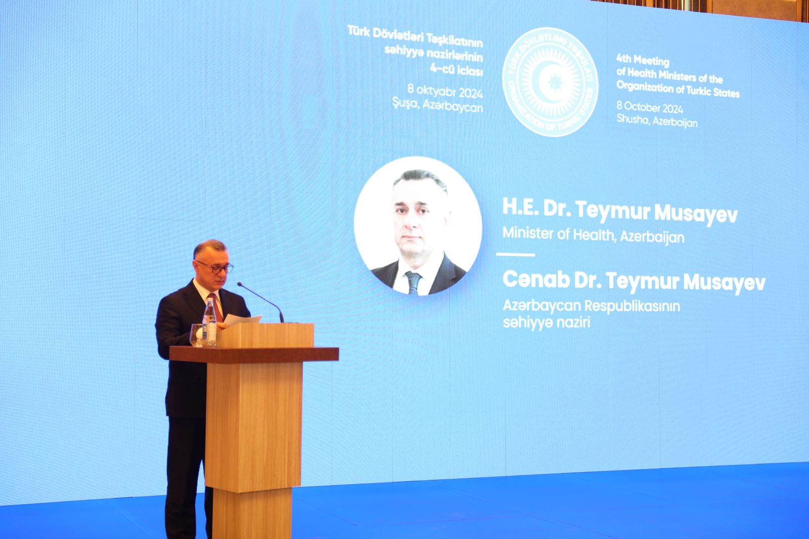 A speaker at a podium addresses an audience, with a large screen displaying information about Dr. Teymur Musayev, Azerbaijan's Minister of Health.