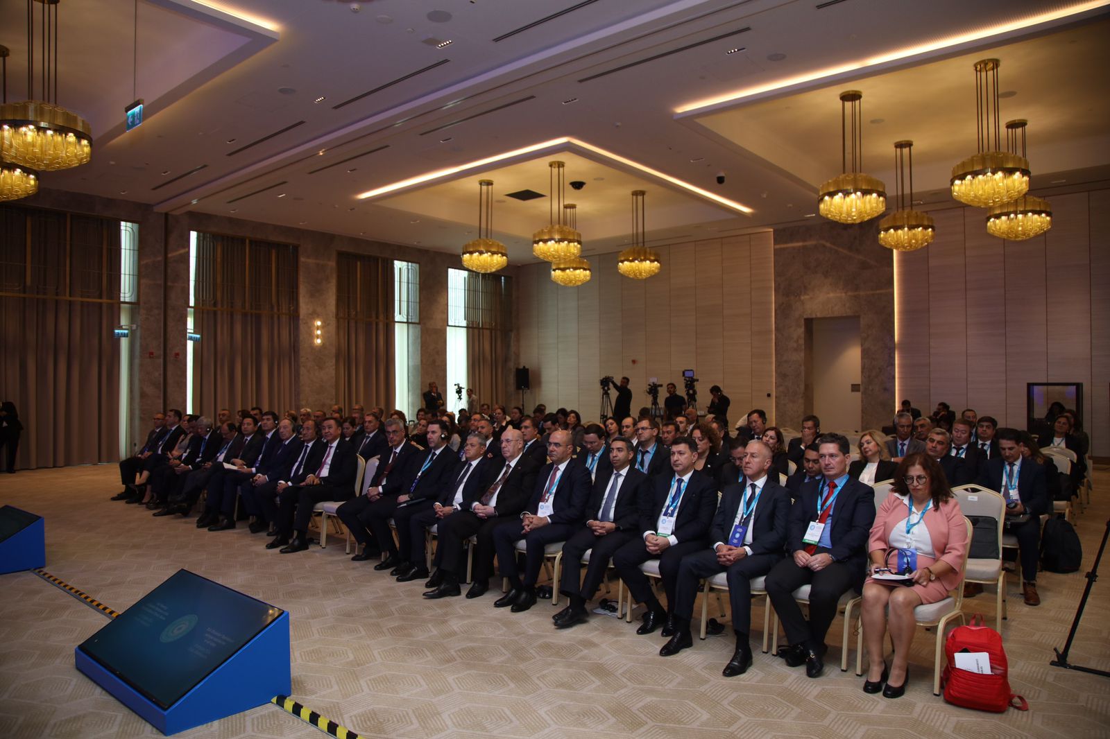 A large audience of professionals seated in a conference room, attentively listening to a presentation. Modern decor and lighting are visible.