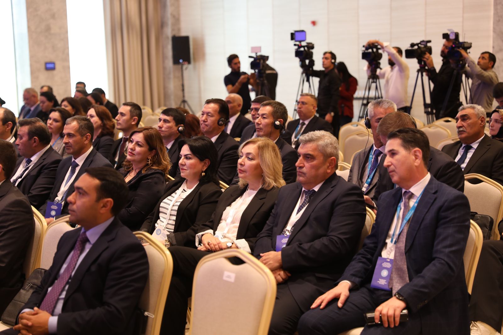 A group of attendees in formal attire sits in a conference setting, with cameras and media personnel in the background.