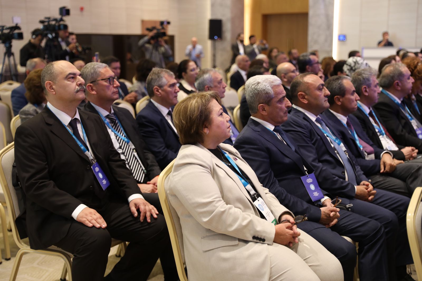 A group of attendees seated in a conference hall, listening attentively. They are dressed in formal attire, with name tags visible.