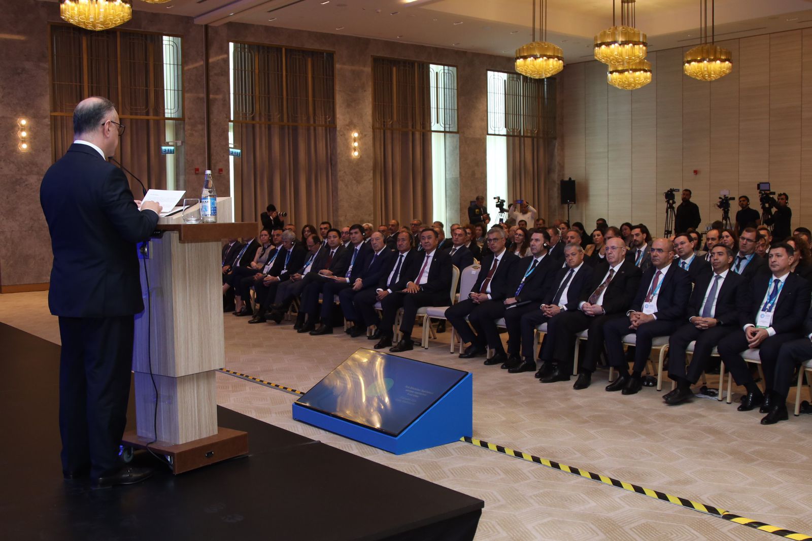 A speaker addresses an audience in a formal setting, with attendees seated and focused on the presentation.