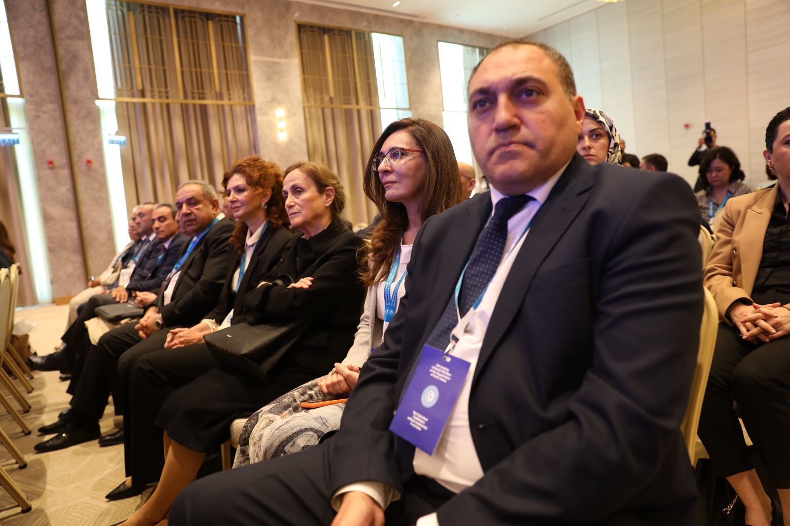 A group of people seated in a conference setting, attentively listening. They are dressed in formal attire.