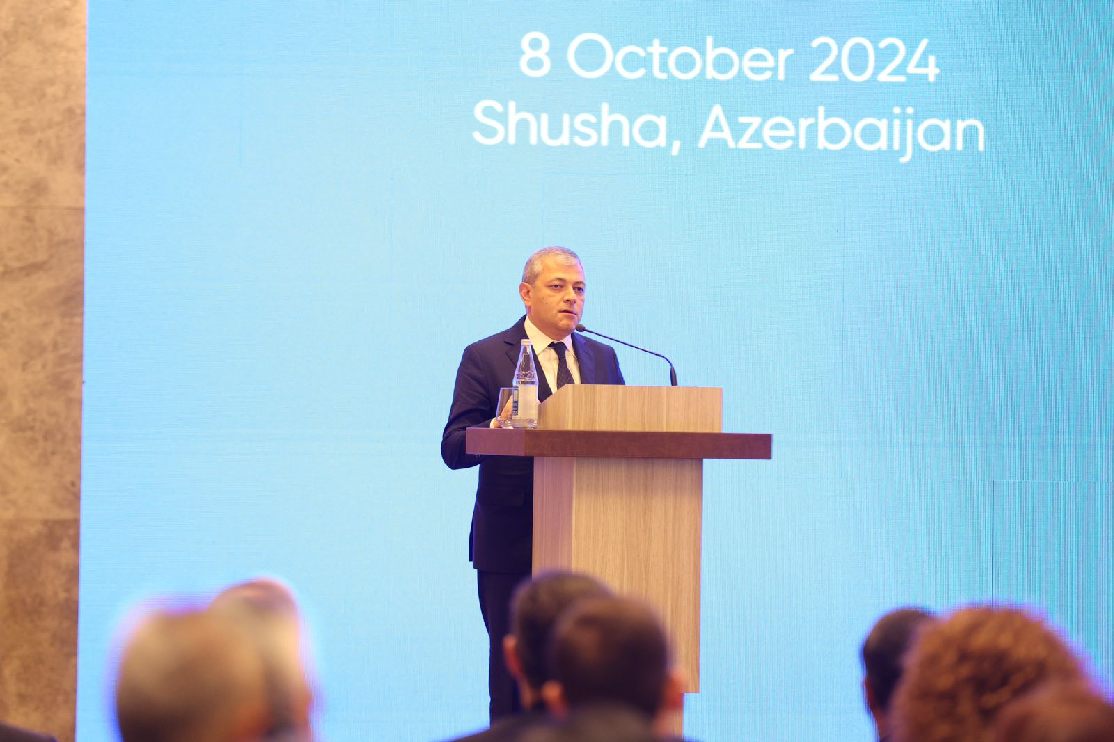 A speaker at a podium addresses an audience, with a blue screen displaying "8 October 2024, Shusha, Azerbaijan" in the background.