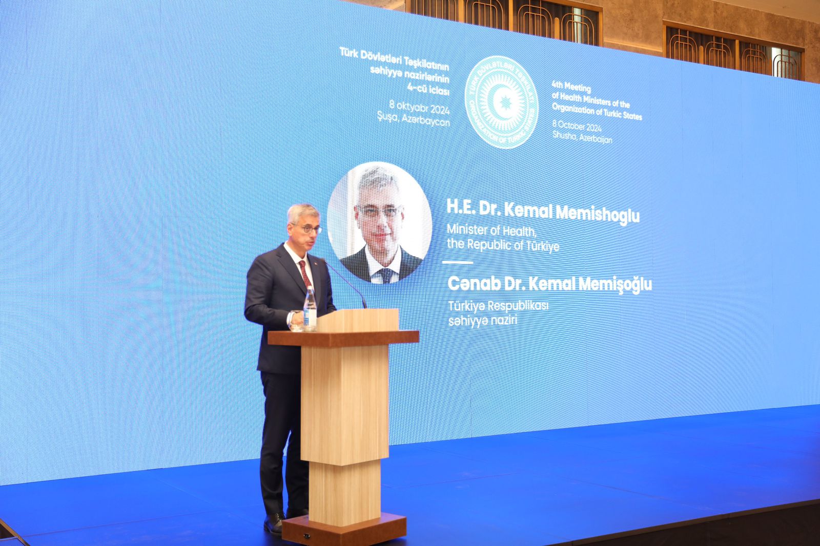 A man stands at a podium speaking at a conference, with a large screen displaying his name and title in the background.