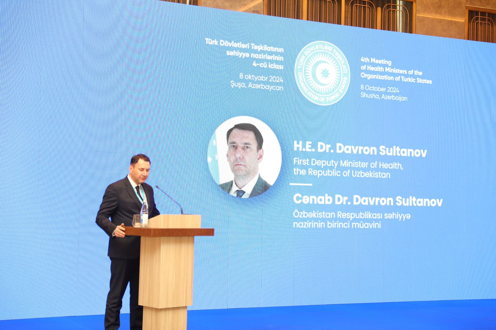 A man in a suit speaks at a podium during a health ministers' meeting, with a large screen displaying his name and title behind him.