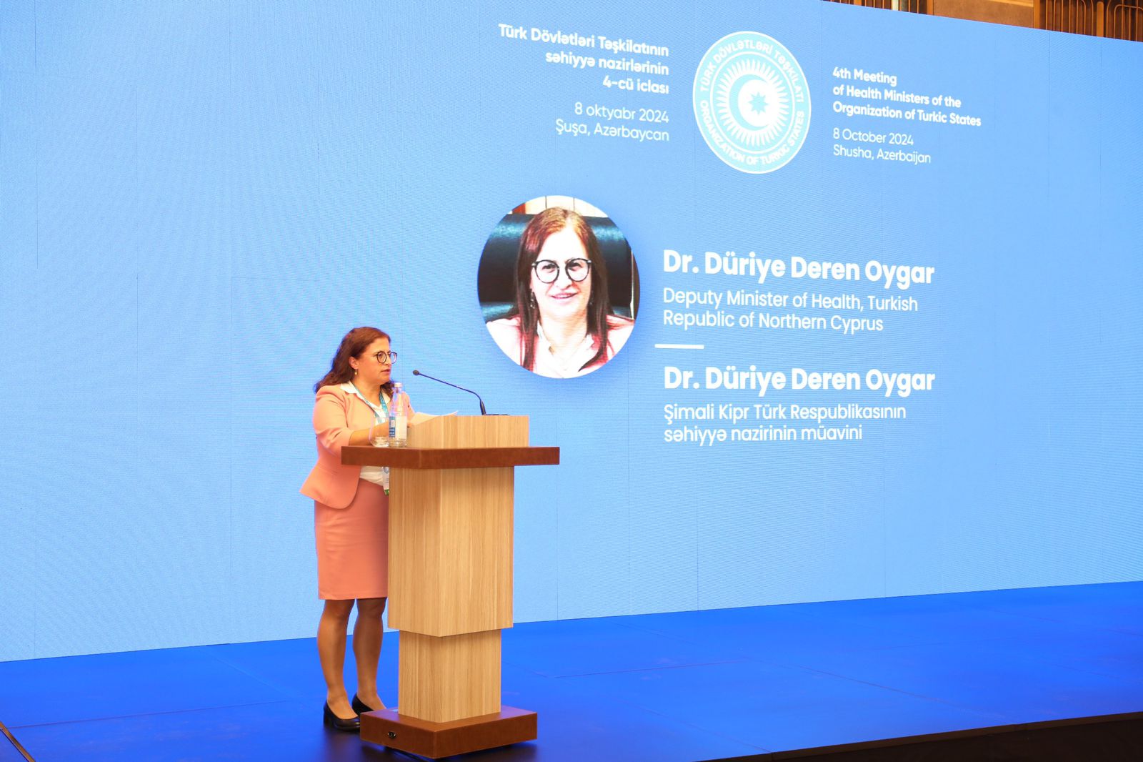 A woman in a pink suit speaks at a podium during a health ministers' meeting, with a large screen displaying her name and title.