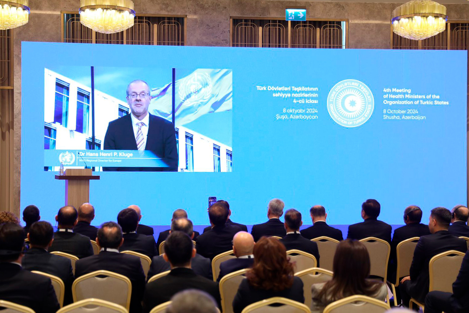 A conference scene with attendees watching a video presentation by Dr. Hans Henri P. Kluge on a large screen.