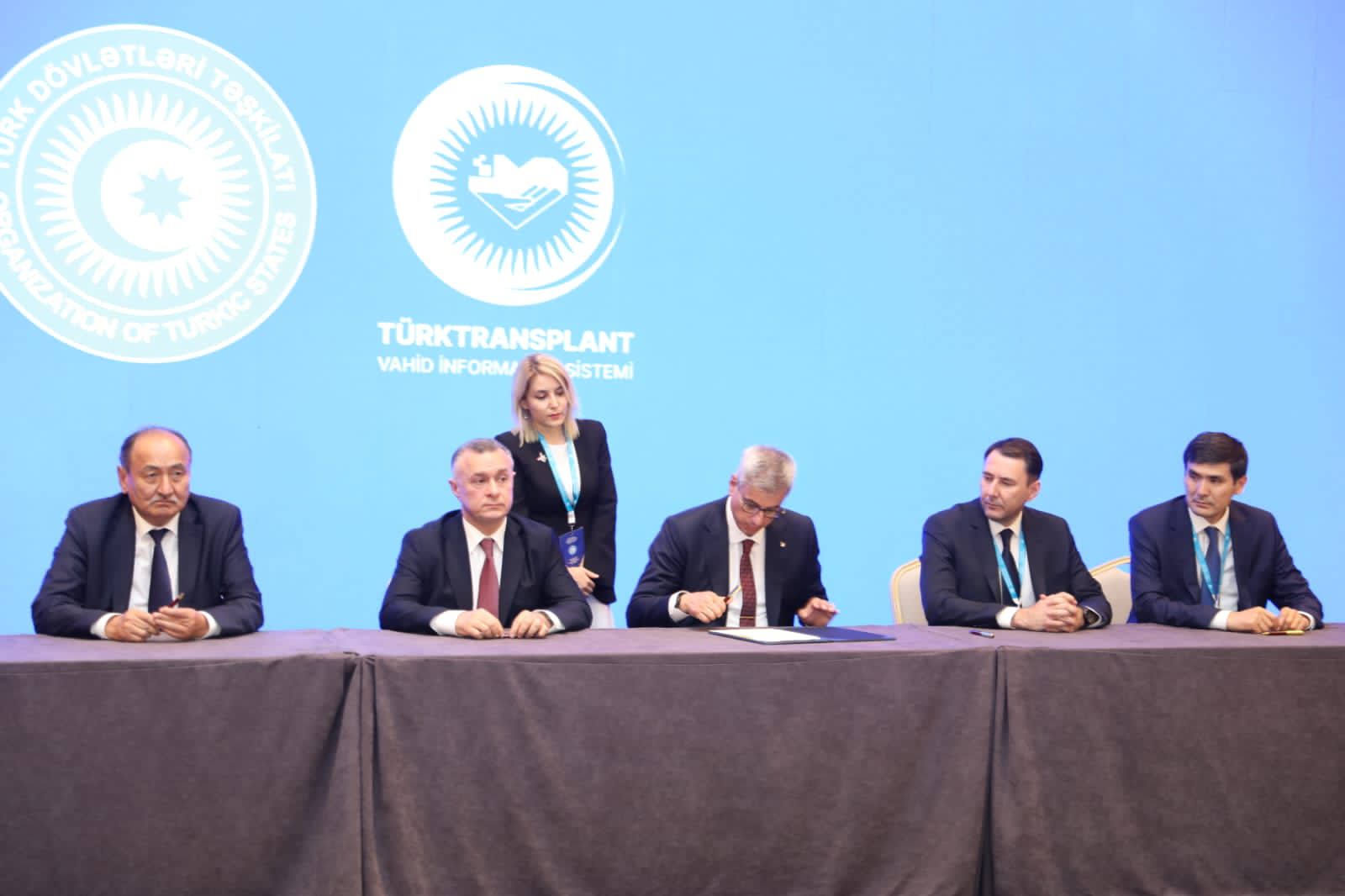 A group of officials seated at a table, with one person signing a document. Background features logos related to Turkic organizations.