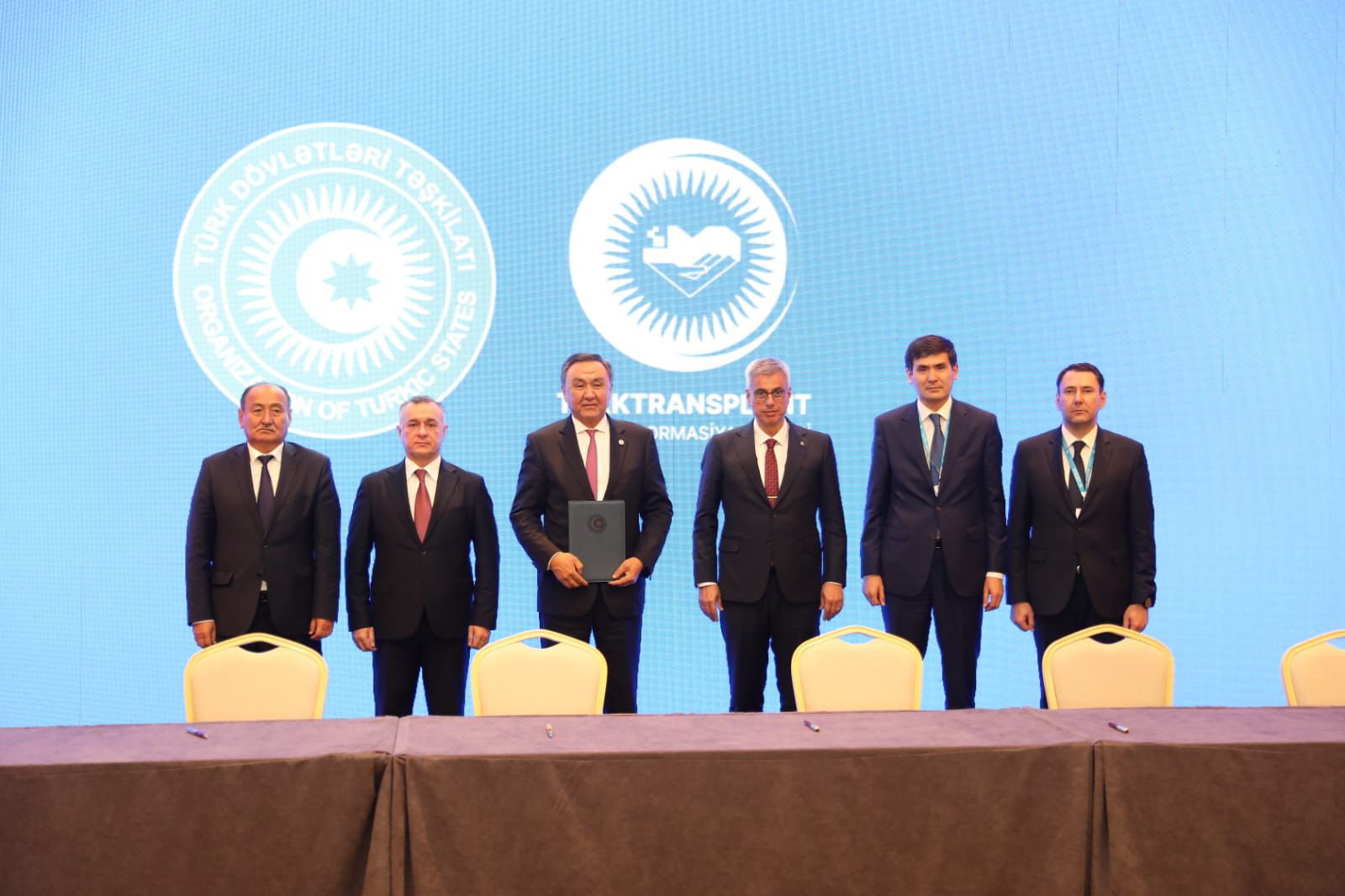 A group of six men in formal attire stands in front of a blue backdrop with organizational logos, with a table in the foreground.