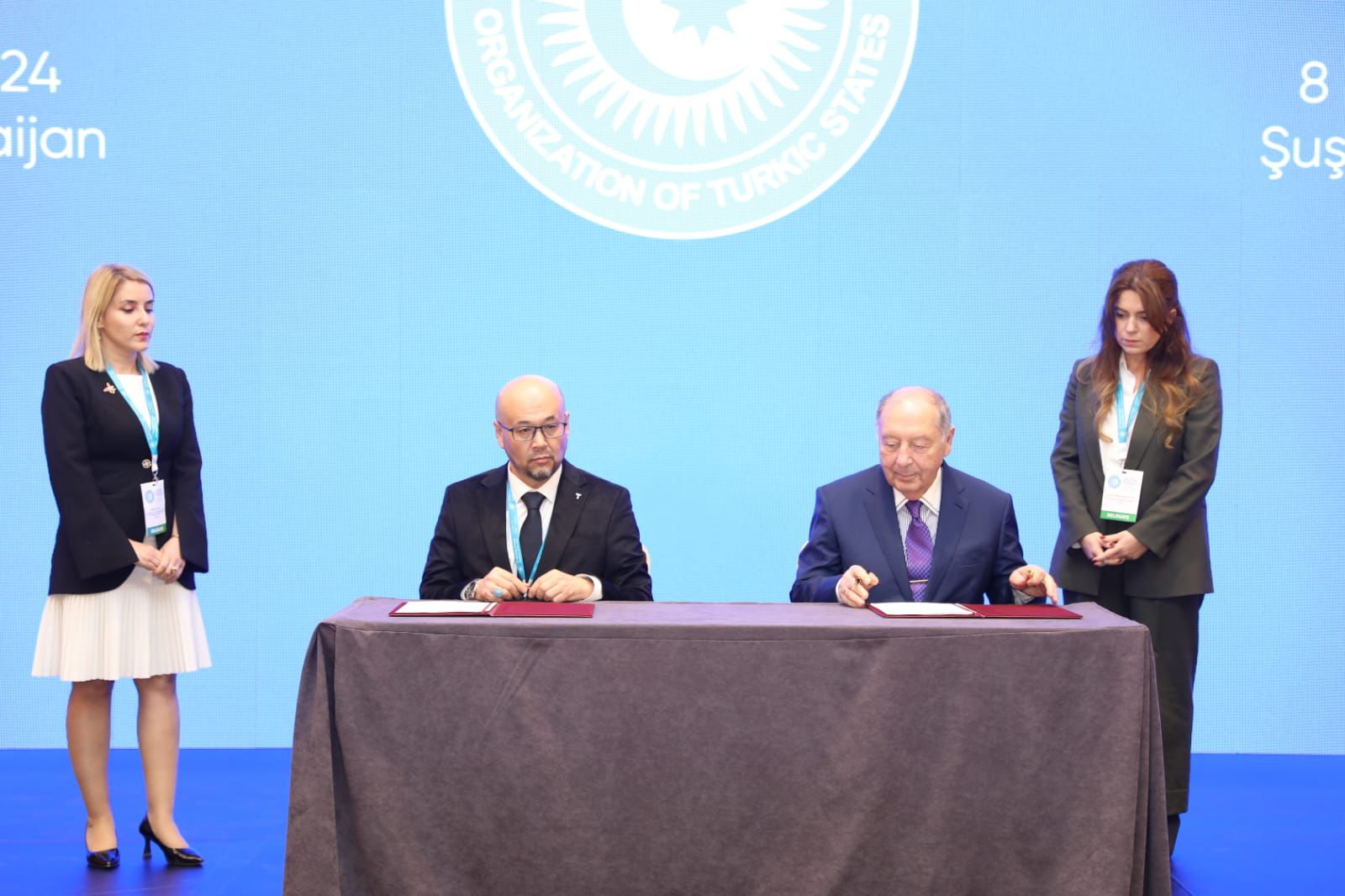 Two men are signing documents at a table, flanked by two women. A blue backdrop displays the "Organization of Turkic States" logo.