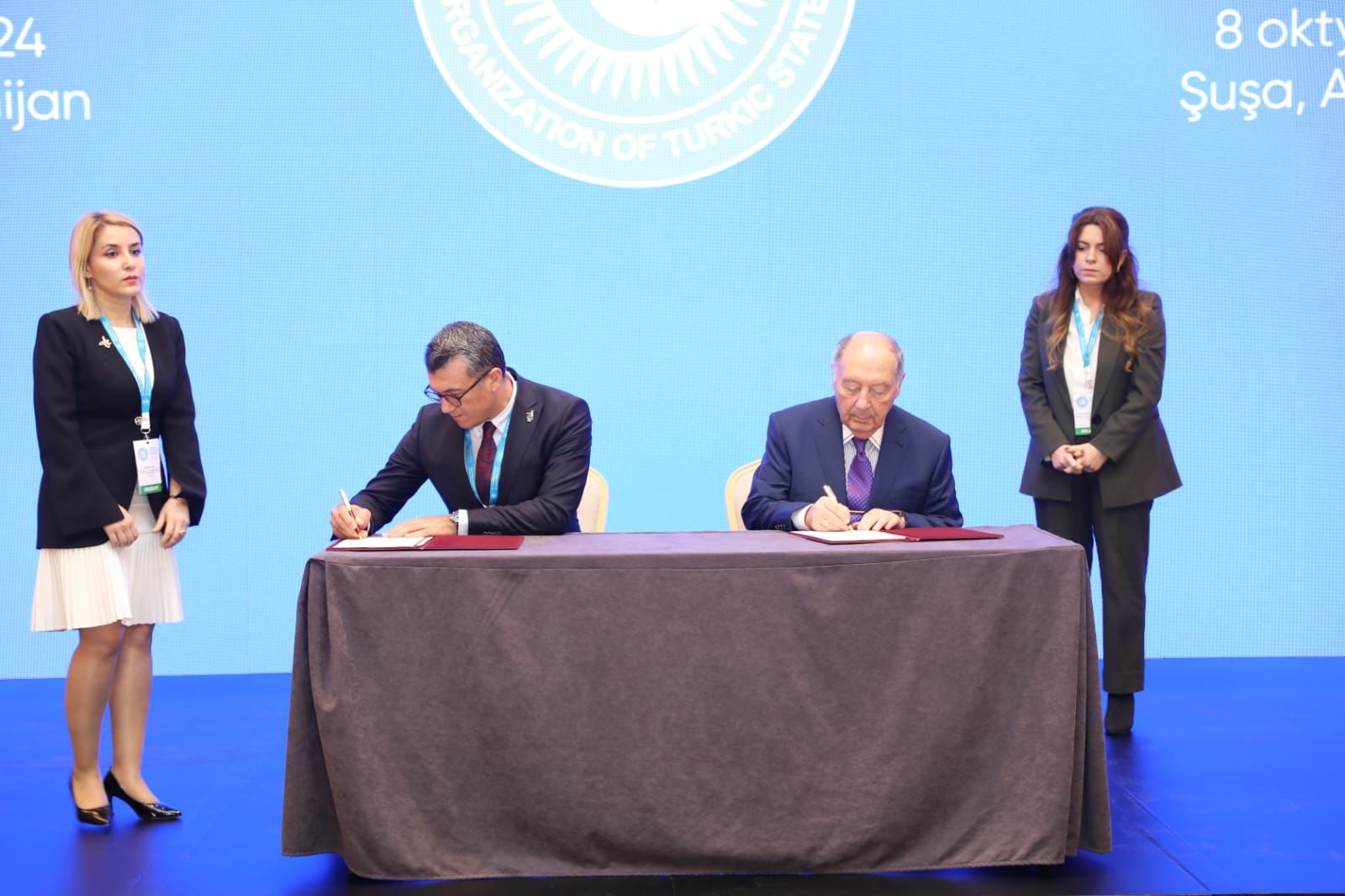 Two men are signing documents at a table, with two women standing nearby. The background features a blue screen with a logo.