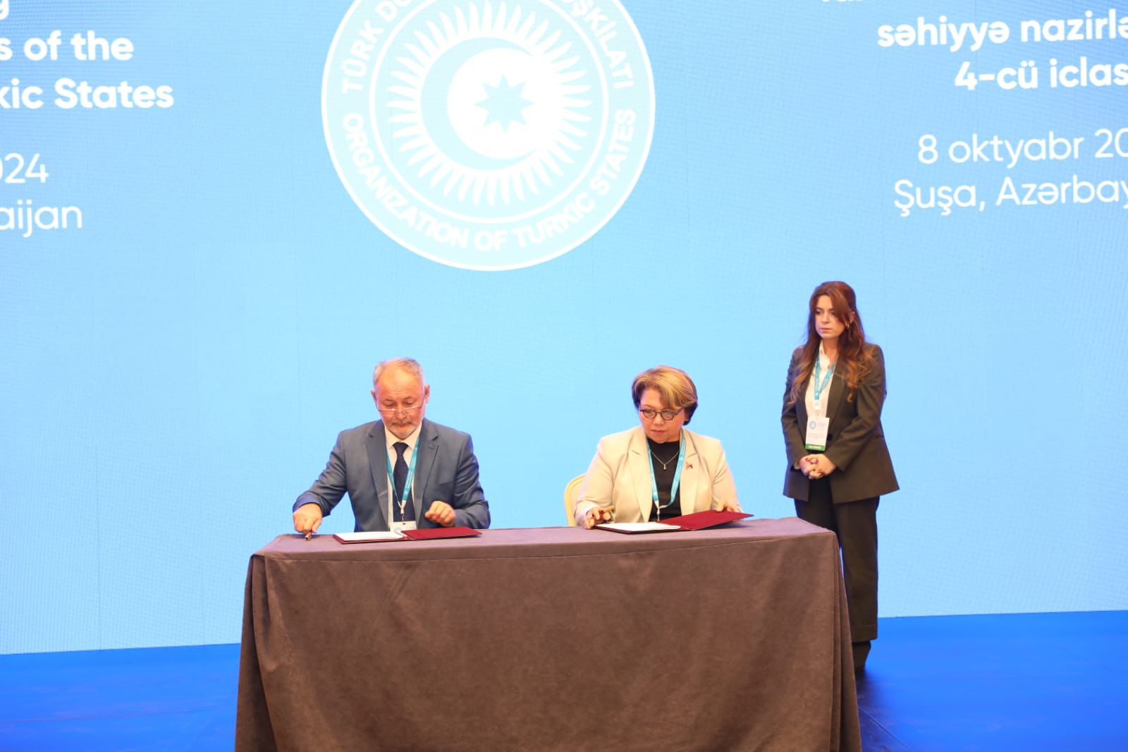 Two officials are signing documents at a table during a conference, with a woman standing behind them. A blue backdrop features the logo of the Turkic States.