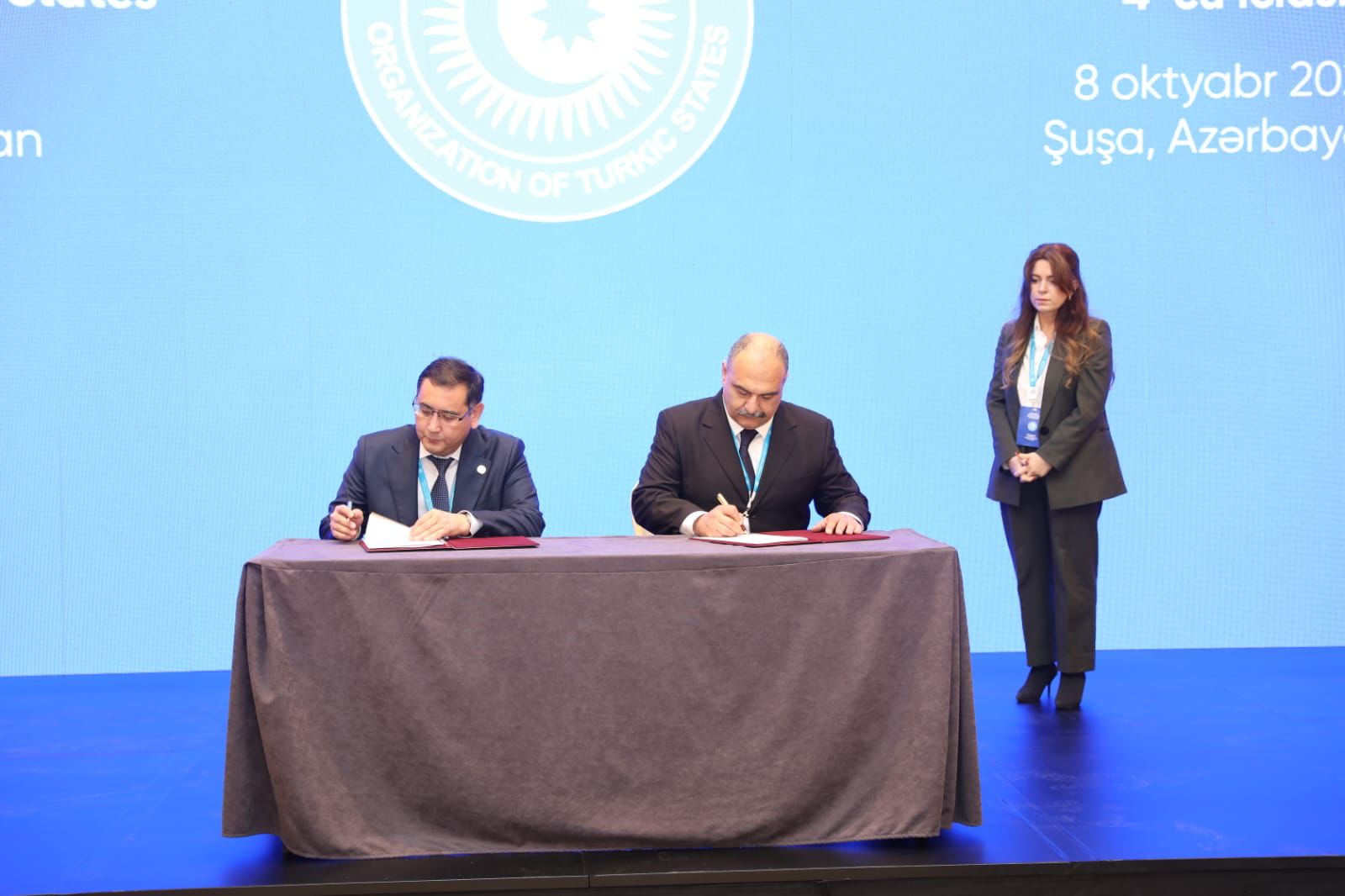 Two officials are signing documents at a table, with a woman standing behind them. The background features a blue screen with a logo.