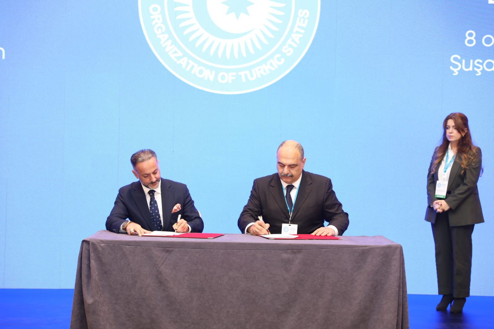 Two men are signing documents at a table, with a woman standing in the background. A blue screen displays the logo of the Organization of Turkic States.