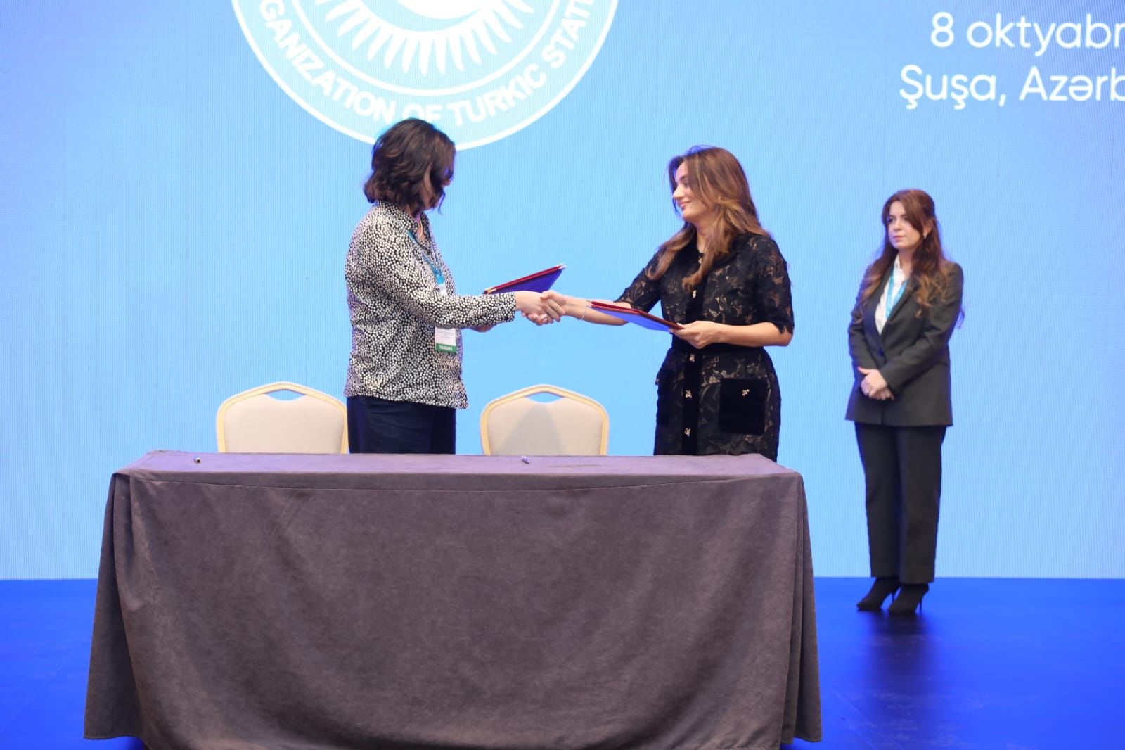 Two women are shaking hands while exchanging documents at a signing event, with a third woman standing in the background.