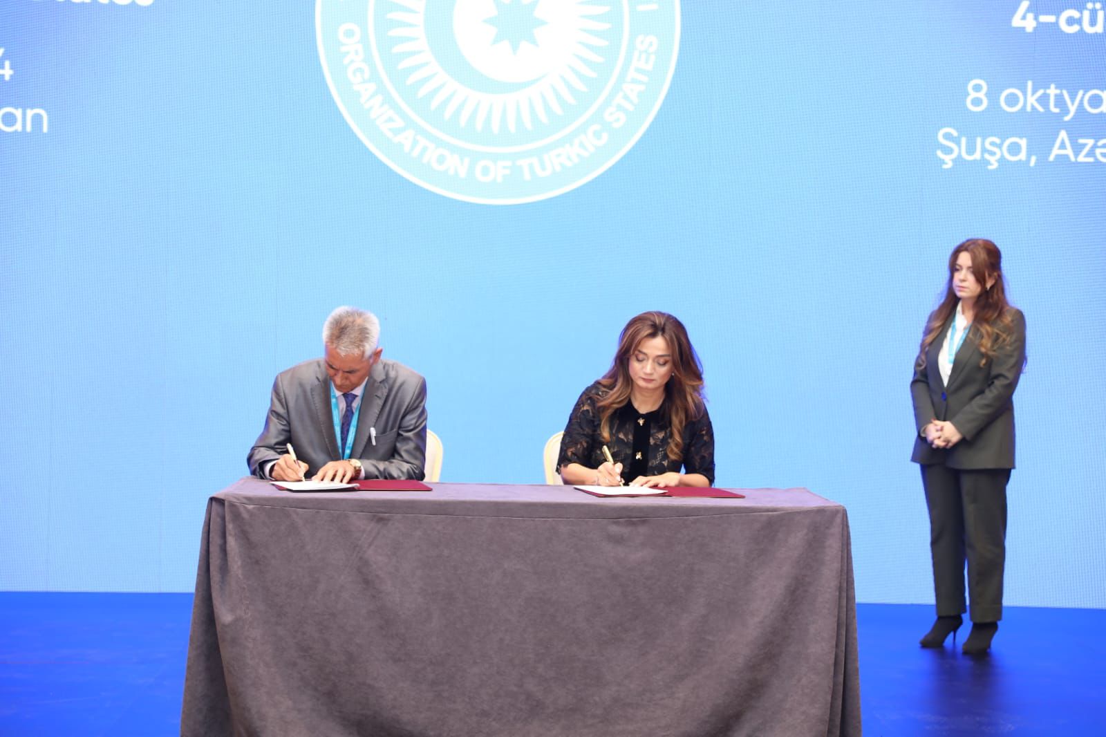 Two individuals are signing documents at a table, with a third person standing behind them. The background features a blue screen with a logo.